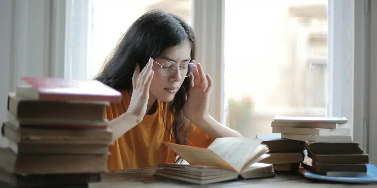 Ragazza con occhiali e mani sulle tempie, circondata da libri. Potrebbe soffrire di mancanza di concentrazione cause.