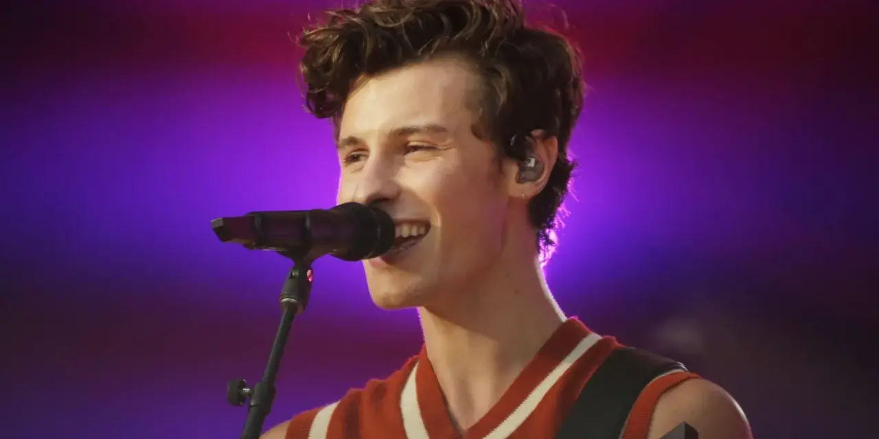 Shawn Mendes śpiewa swoje utwory na koncercie, z mikrofonem przed ustami i gitarą na ramieniu.