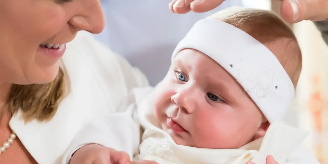 Uśmiechnięta mama trzyma dziecko w białej szatce. Dłoń ojca błogosławi malucha. Zastanawiasz się, ile kosztuje chrzest?
