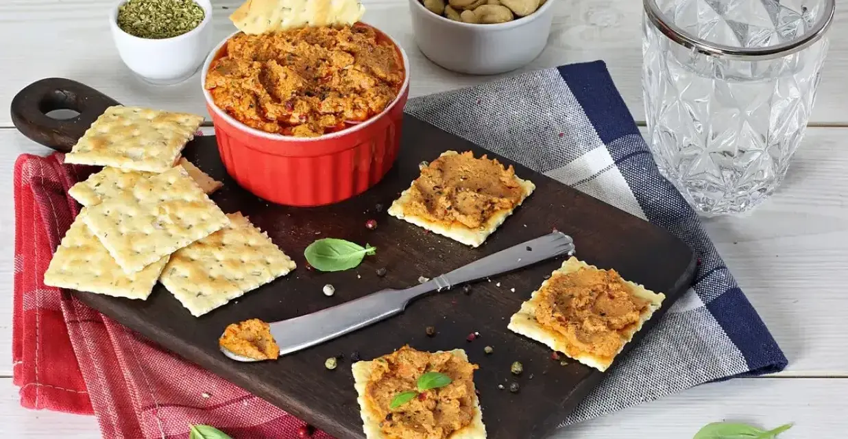 Pyszna pasta z suszonych pomidorów na krakersach, obok miseczka orzechów i szklanka wody. Idealna przekąska!