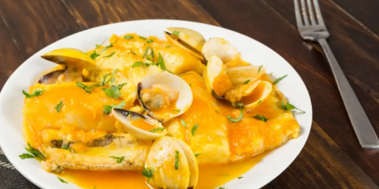 Merluza a la marinera con almejas, servida en un plato blanco sobre una mesa de madera oscura.