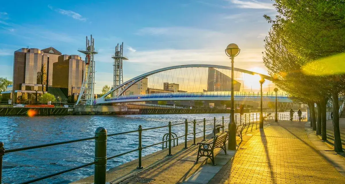 Promenade ensoleillée au bord de l'eau, avec un pont moderne et des bâtiments reflétant le soleil. Idéal pour visiter Manchester ou Liverpool.