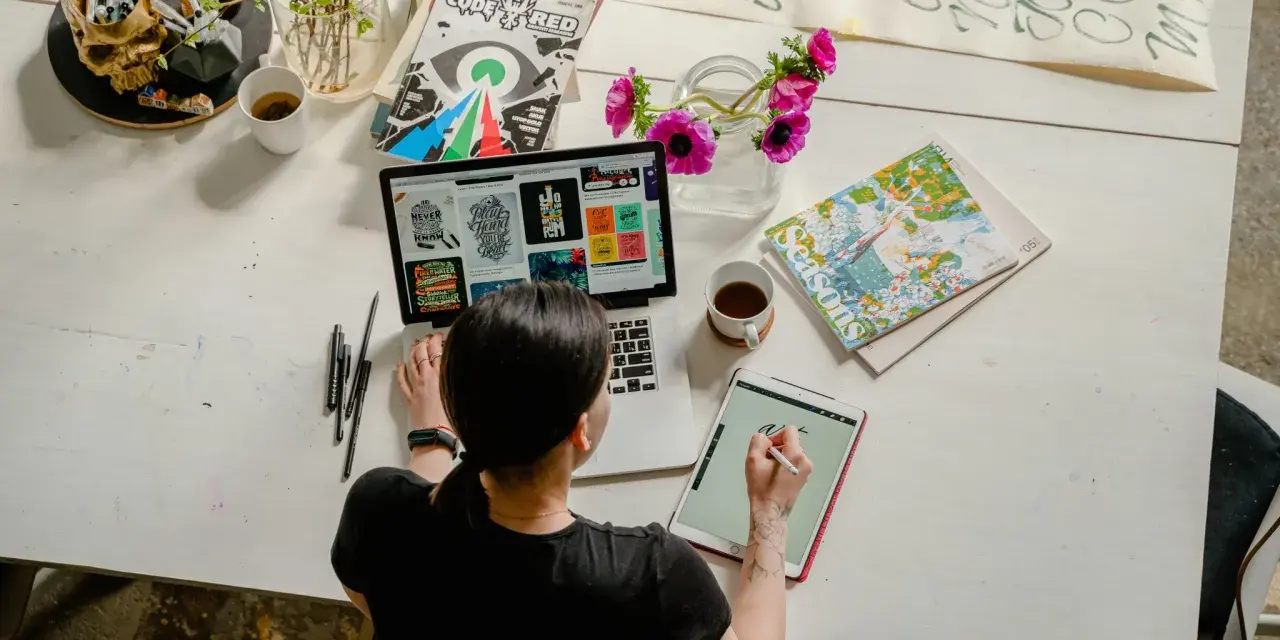 Kobieta pracuje online w domu, projektując na laptopie i tablecie. Na biurku kwiaty, kubek kawy i czaszki z pisakami.