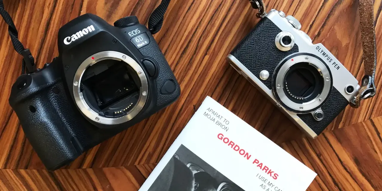 Dwa aparaty, Canon EOS 6D Mark II i Olympus PEN, leżą obok książki Gordona Parks'a "Aparat to moja broń". Pełna klatka i mniejszy aparat gotowe do akcji.