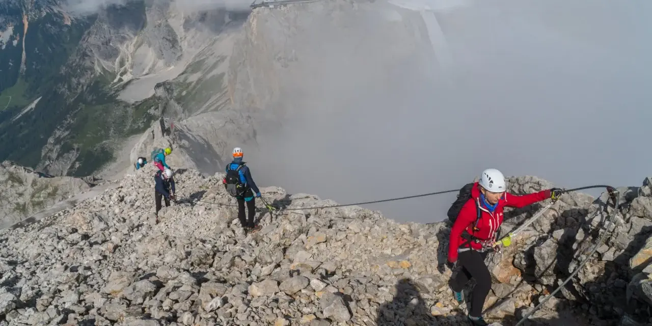 Grupa turystów pokonuje via ferrata polska. Wspinają się po skalistym grzbiecie, asekurowani linami. W tle widać góry i budynek na szczycie.