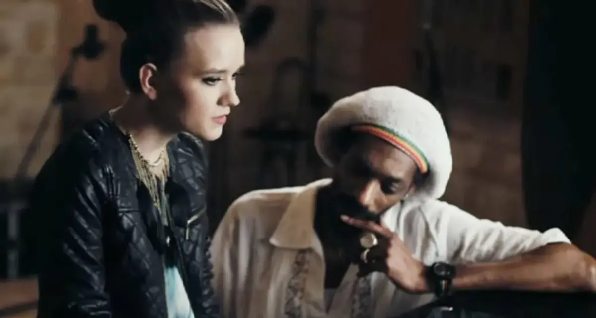 Snoop Dogg w białej koszuli i beretce Rasta, rozmawiający z młodą kobietą. Może to być scena z jednego z jego **snoop dogg utworów**.