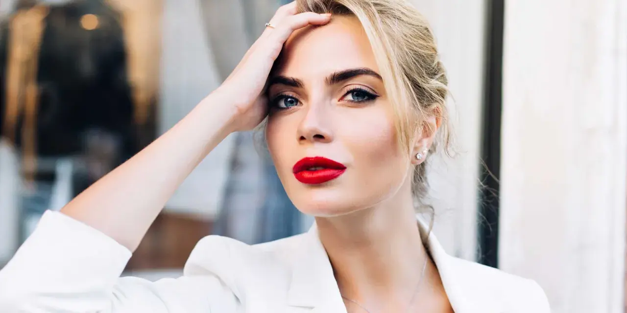 Frau mit roten Lippen und weißem Blazer, Hand am Kopf.