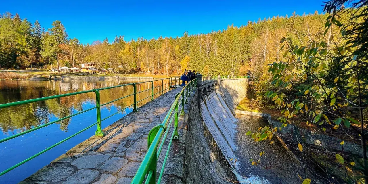 Kamienna zapora z zieloną balustradą, wodospadem i jesiennym lasem w tle.
