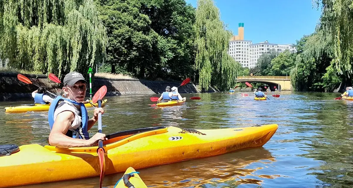 Eine Gruppe von Menschen paddelt auf gelben Kajaks auf einem Fluss in Berlin. Bäume säumen das Ufer, im Hintergrund ist ein Gebäude zu sehen.