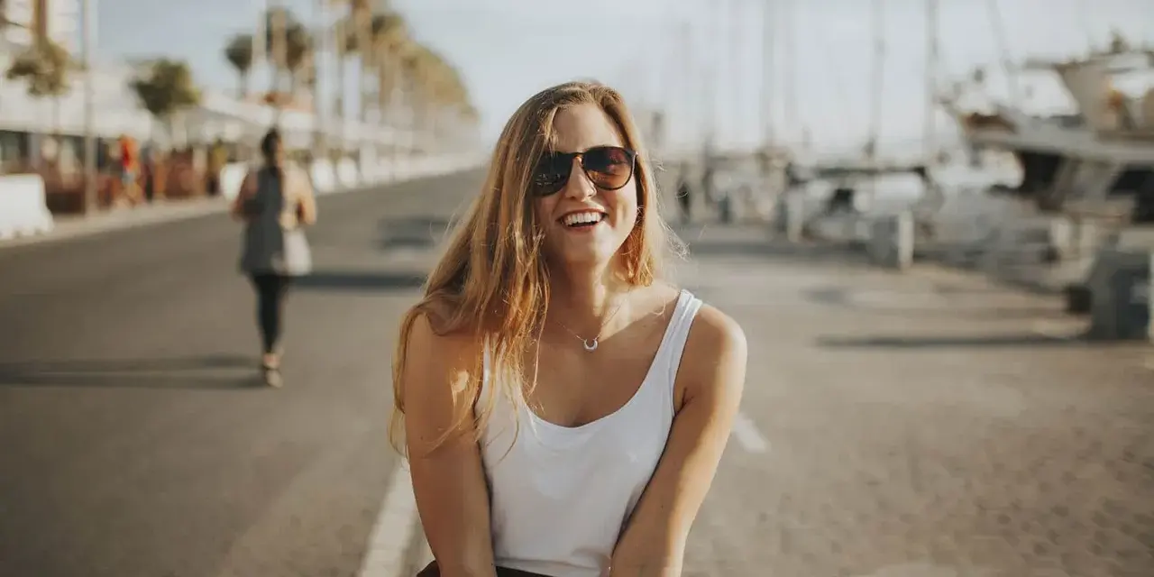 Glückliche junge Frau mit Sonnenbrille und weißem Top genießt ihr Praktikum in Spanien am Hafen.