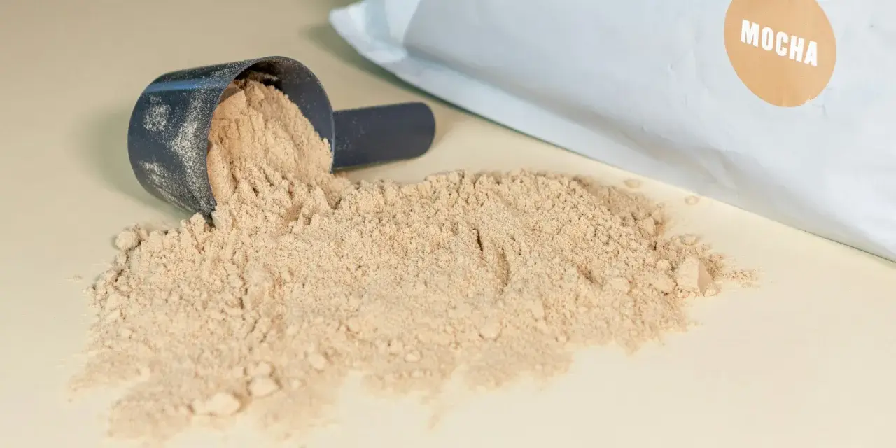 Moccha-smakande proteinpulver spillt från en skopa, med påsen i bakgrunden. Perfekt för att hålla koll på proteinpulver kcal.