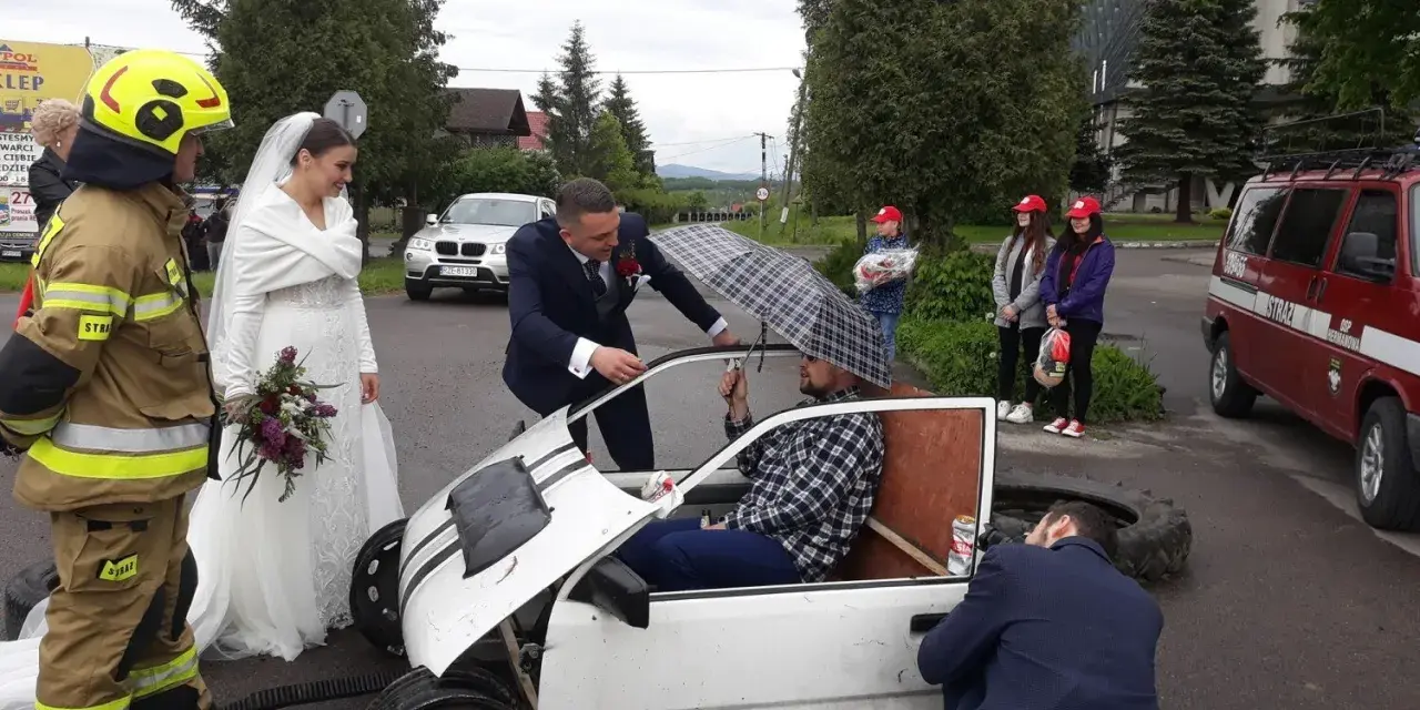 Brama weselna z udziałem strażaka, pana młodego i panny młodej. Pan młody rozmawia z druhem siedzącym w prowizorycznym samochodzie.