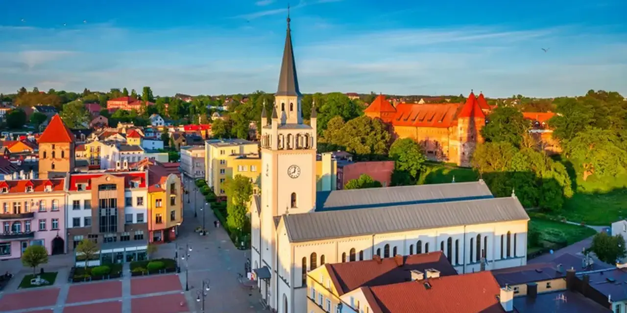 Słoneczny rynek w Bytowie z kościołem i zamkiem w tle. Kolorowe kamienice tworzą urokliwy krajobraz.