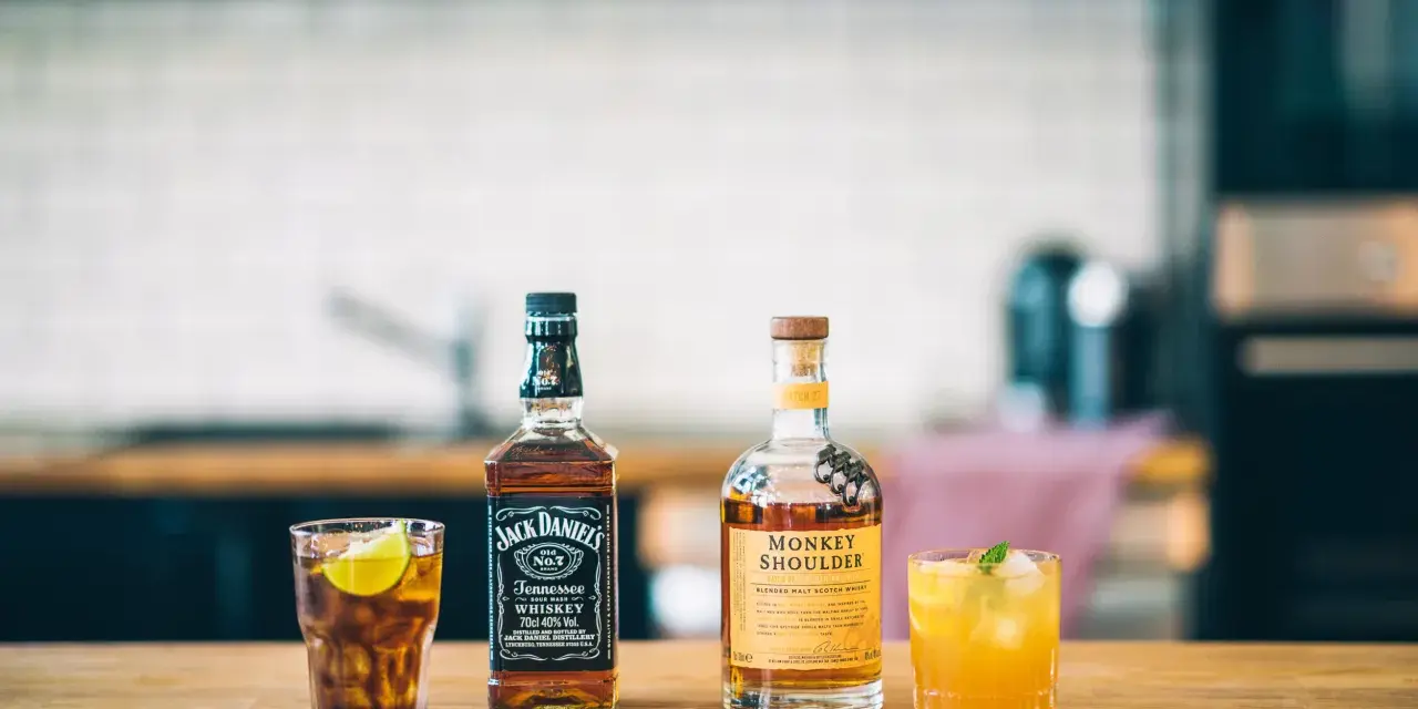 Zwei Flaschen Whiskey, Jack Daniel's und Monkey Shoulder, stehen neben zwei Cocktails auf einem Holztisch.