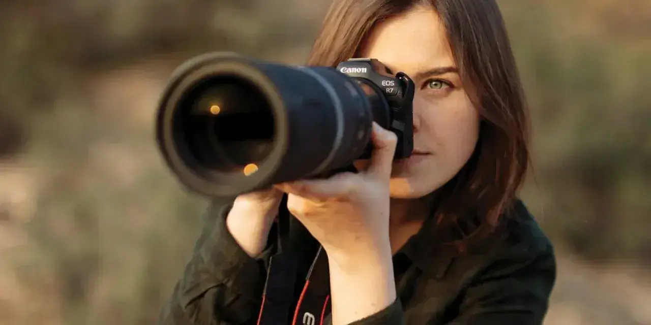 Kobieta z aparatem Canon EOS R7, skupiona na kadrze. Jej oczy błyszczą, a obiektyw sugeruje zaawansowane techniki fotograficzne.