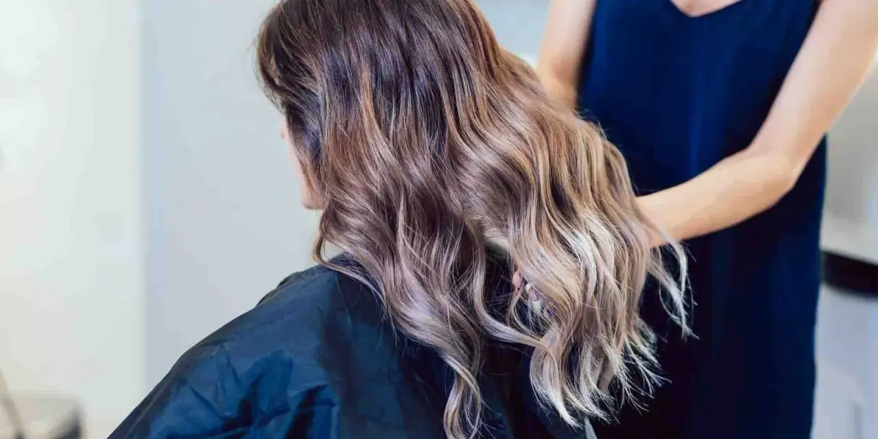 Kobieta z falowanymi włosami w odcieniach brązu i subtelnym balejażem blond. Fryzjerka poprawia jej fryzurę.