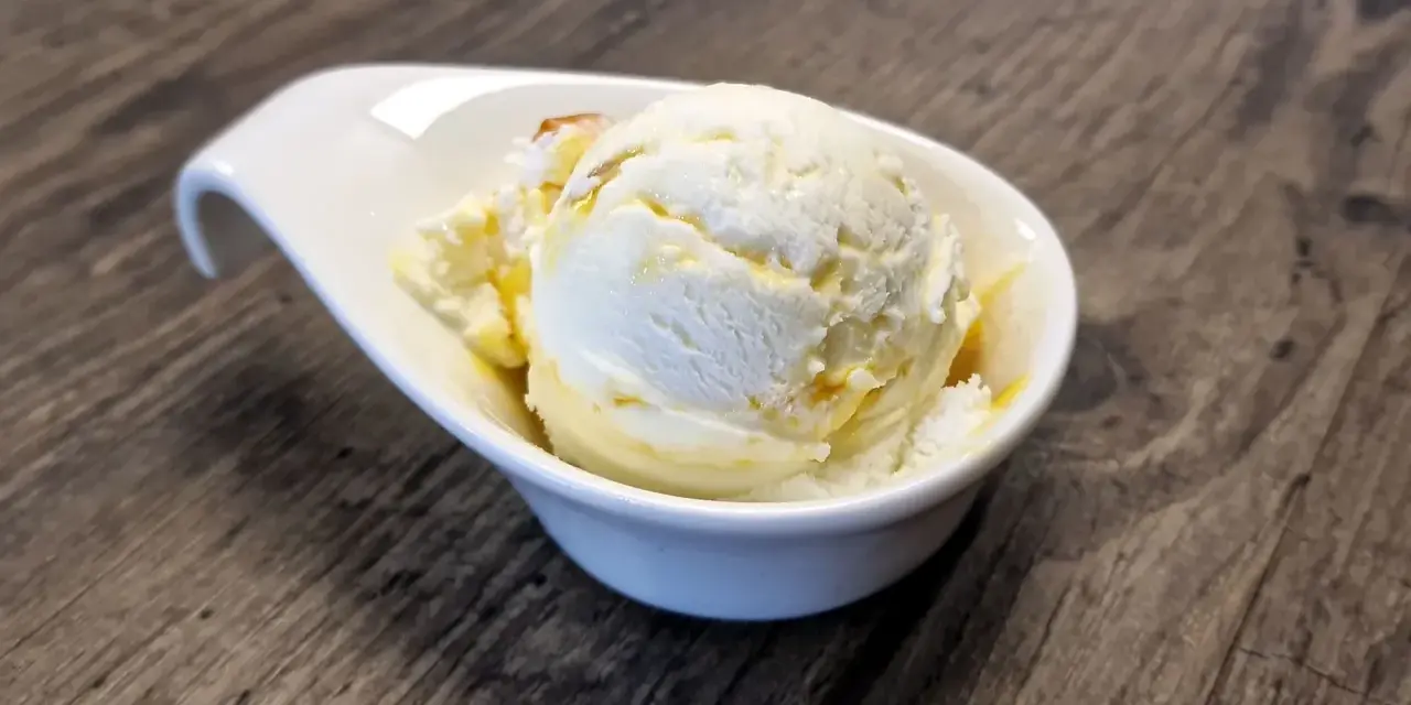 Ein Löffel cremiges Buttermilch-Eis, beträufelt mit Honig, in einer weißen Schale auf Holztisch.