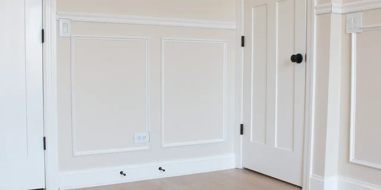 A room with a DIY chair rail, wainscoting, and white doors. The walls are painted a light cream color.