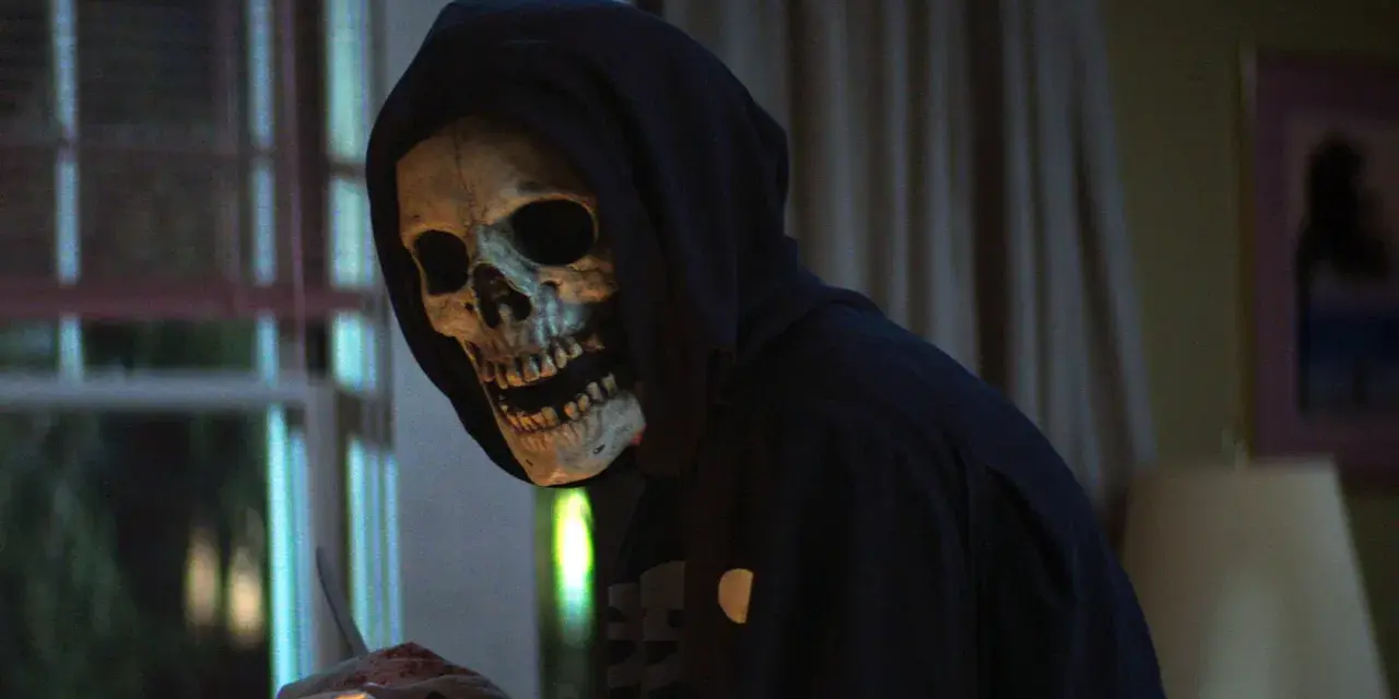 Eine Person mit Totenkopfmaske und Kapuze hält blutige Gegenstände.
