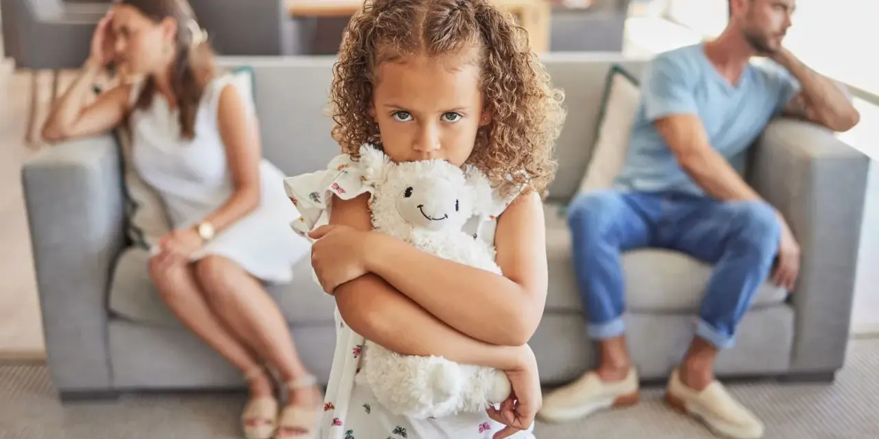 Ein Kind umklammert sein Stofftier, während die Eltern im Hintergrund getrennt sitzen. Ein Bild, das die emotionale Belastung eines Trennungsjahres mit Kindern widerspiegelt.