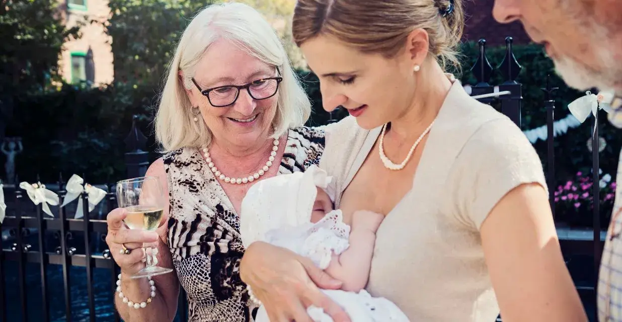 Elegancka mama karmi dziecko na chrzcinach. Babcia z kieliszkiem wina podziwia wnuka. Jak się ubrać na chrzciny? Klasycznie i z klasą.