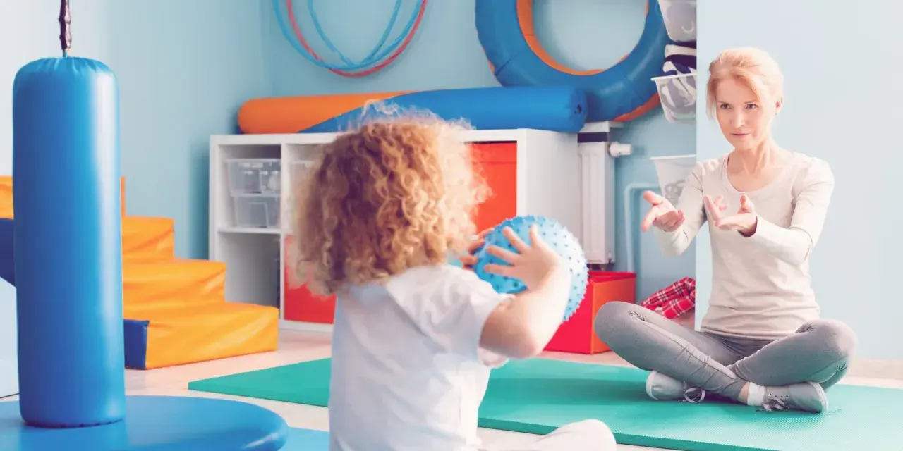 Ergotherapie für Kinder: Eine Therapeutin erklärt einem Kind mit Locken, wie man einen Ball wirft.