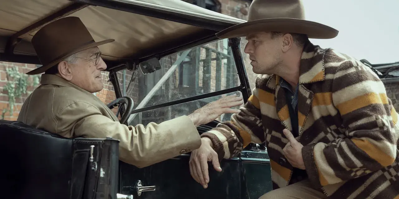 Due uomini in auto discutono animatamente. Uno, al volante, con cappello e giacca chiara, sembra implorare l'altro. Quest'ultimo, con un cappello simile e una giacca a righe, ascolta con espressione tesa, forse coinvolto negli eventi del "killer of the...