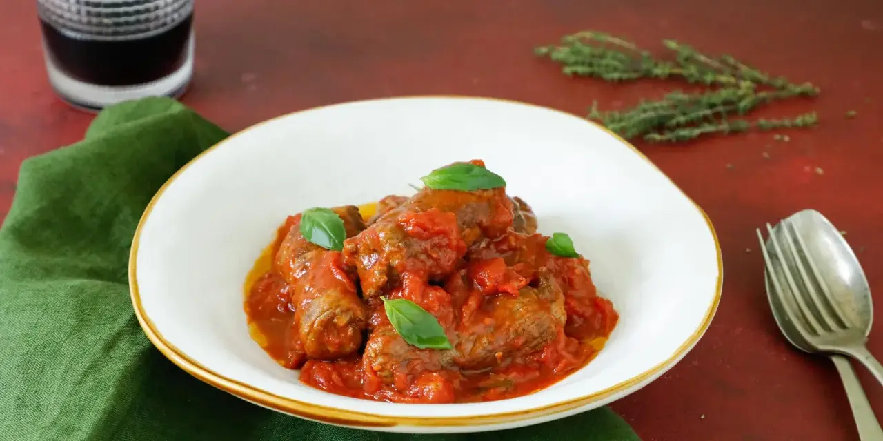 Involtini di carne in padella con sughetto, guarniti con basilico fresco, serviti in un piatto bianco su un tovagliolo verde.