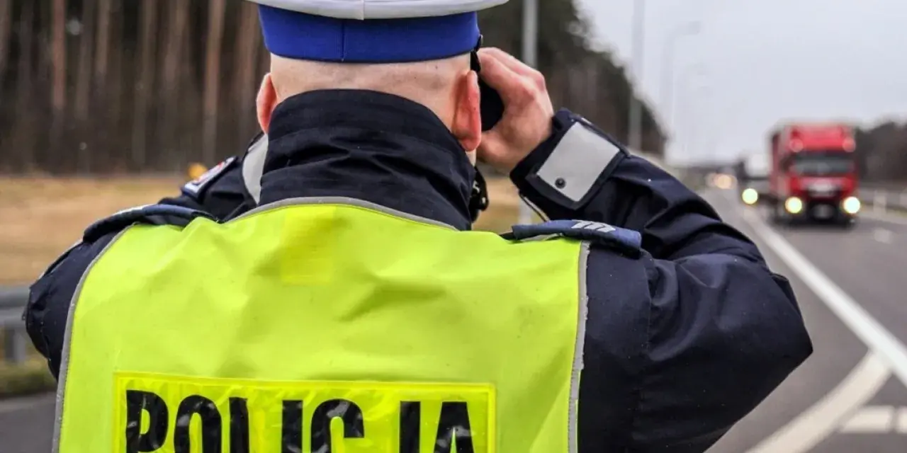Policjant w kamizelce odblaskowej obserwuje drogę, gdzie widać ciężarówkę. Na drodze obowiązuje zakaz wyprzedzania dla ciężarówek.