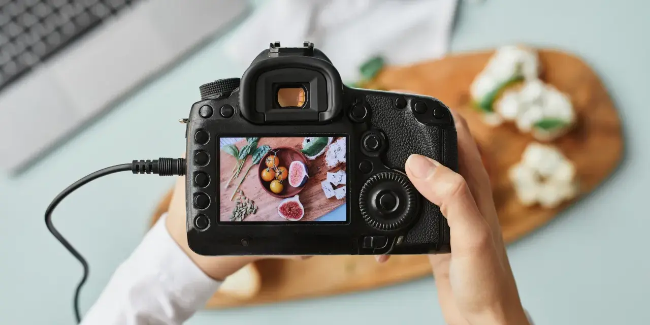 Dłonie trzymające aparat fotograficzny, na którego ekranie widać zdjęcie jedzenia.