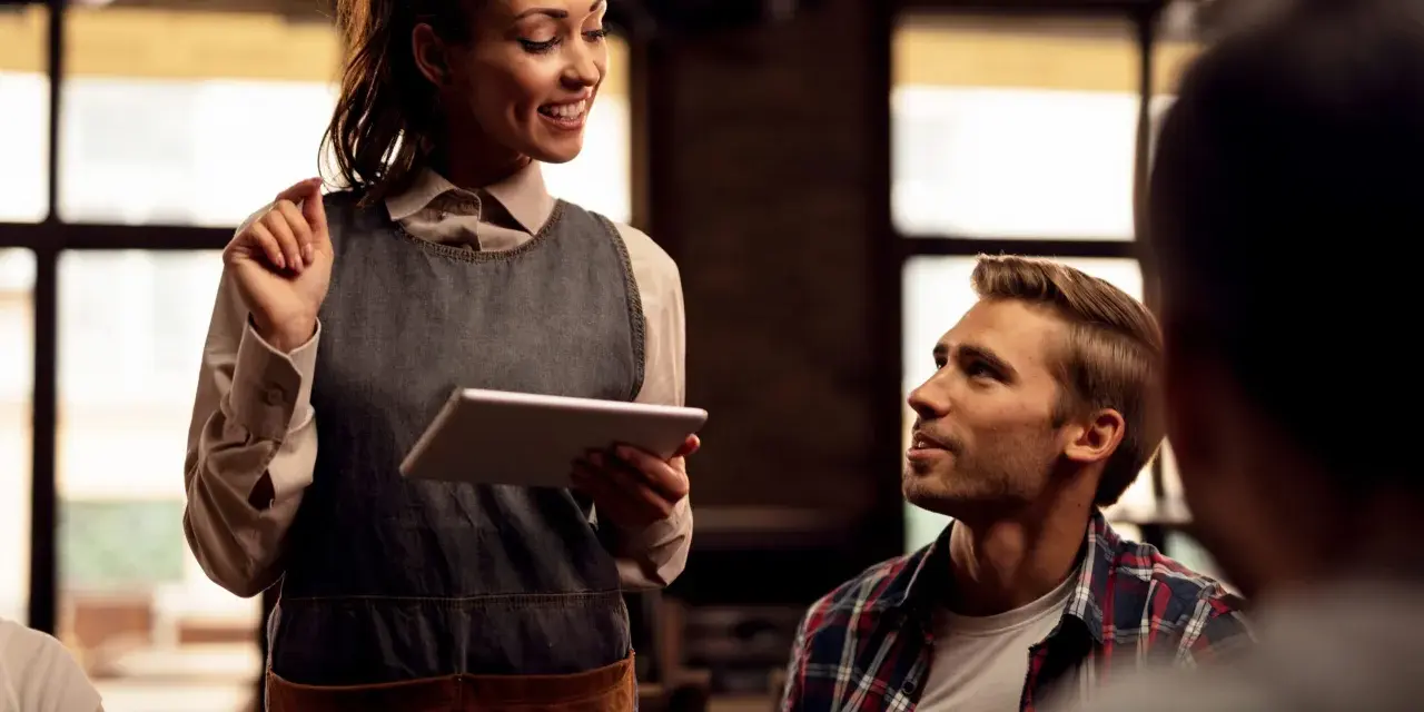 Camarera sonriente atiende a clientes de restaurante, tomando nota en una tablet.