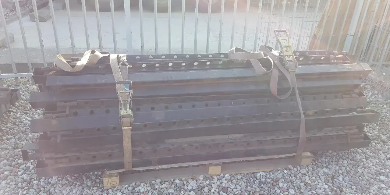 Stos czarnych, metalowych elementów naczepy z pasami transportowymi.