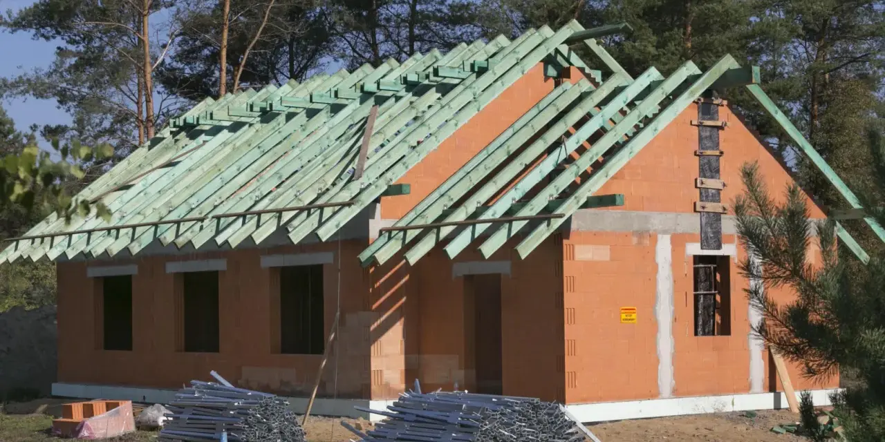 Budowa domu w metodzie gospodarczej: wzniesione ściany z pustaków ceramicznych, gotowy szkielet dachu z zielonych krokwi, materiały budowlane na placu.
