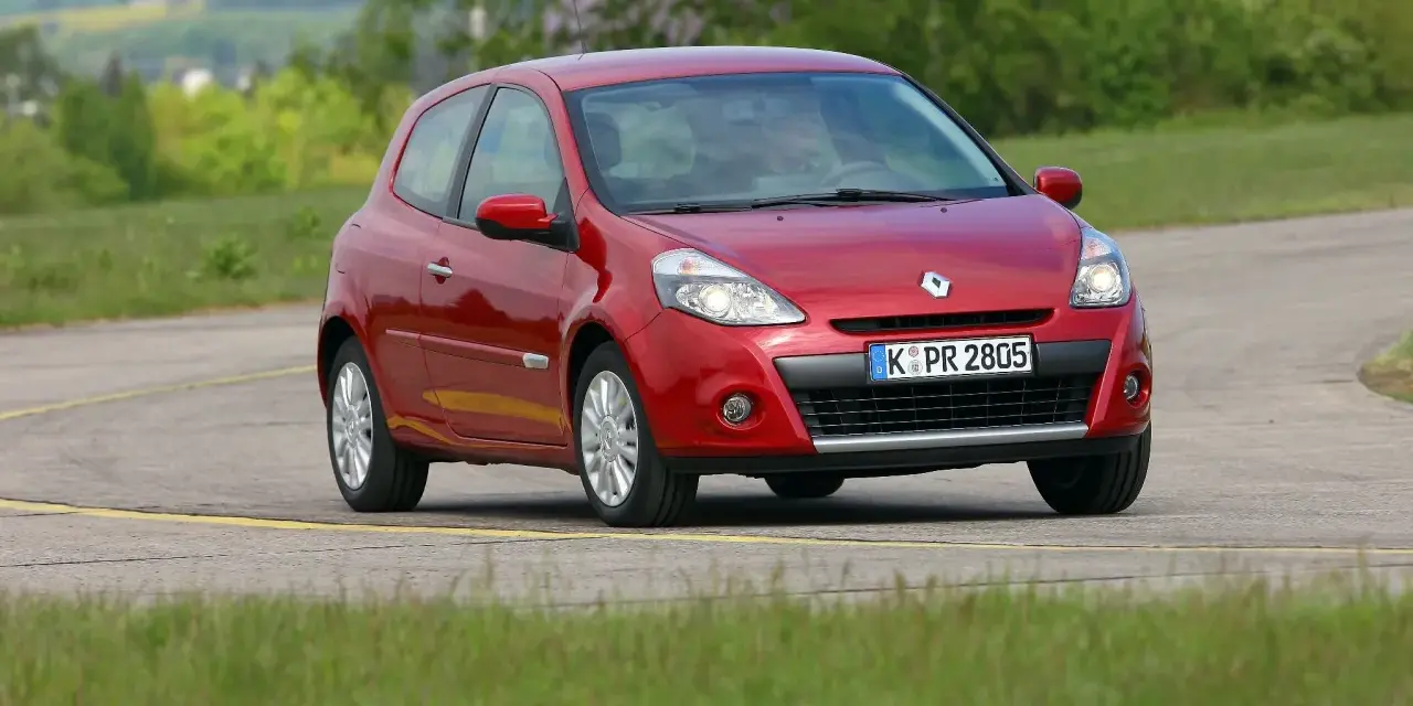 Czerwony Clio 3 na zakręcie drogi, otoczony zielenią.