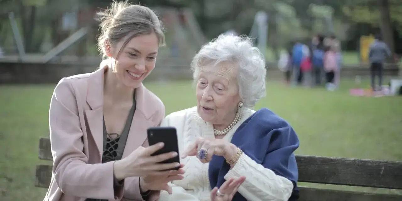 Młoda kobieta pokazuje coś na telefonie starszej pani. To scena z życia, jak z kursu opiekuna osoby starszej, gdzie uczymy się wspierać seniorów w codzienności.