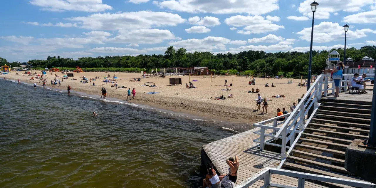Słoneczny dzień nad morzem bałtyckim. Ludzie odpoczywają na piaszczystej plaży, niektórzy kąpią się w wodzie. Drewniane molo prowadzi na plażę.
