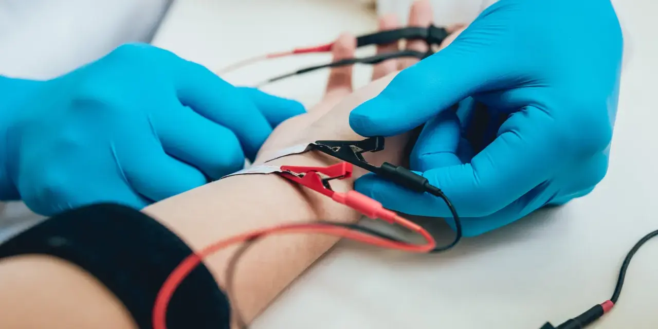 Lekarz w niebieskich rękawiczkach podłącza elektrody do ręki pacjenta. To badanie EMG, które pozwala ocenić funkcje nerwów i mięśni.