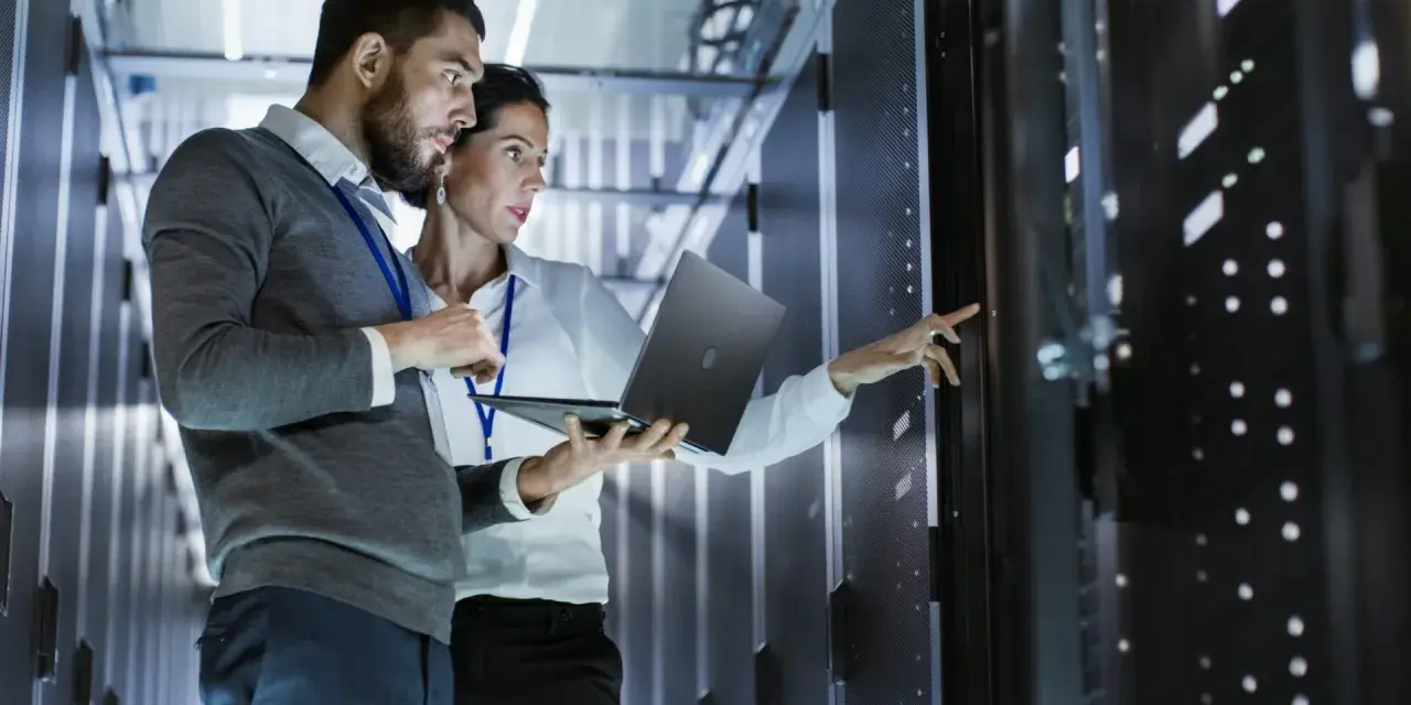 Two IT professionals collaborate on network management, one holding a laptop while the other points to a server rack.