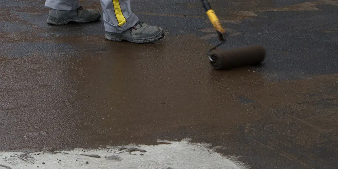 Pracownik wykonuje hydroizolację chudziaka, nakładając masę bitumiczną wałkiem.