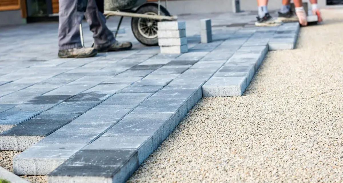 Układanie płyt betonowych na podjeździe – alternatywa dla kostki. Pracownicy budowlani układają szare płyty na podsypce żwirowej.