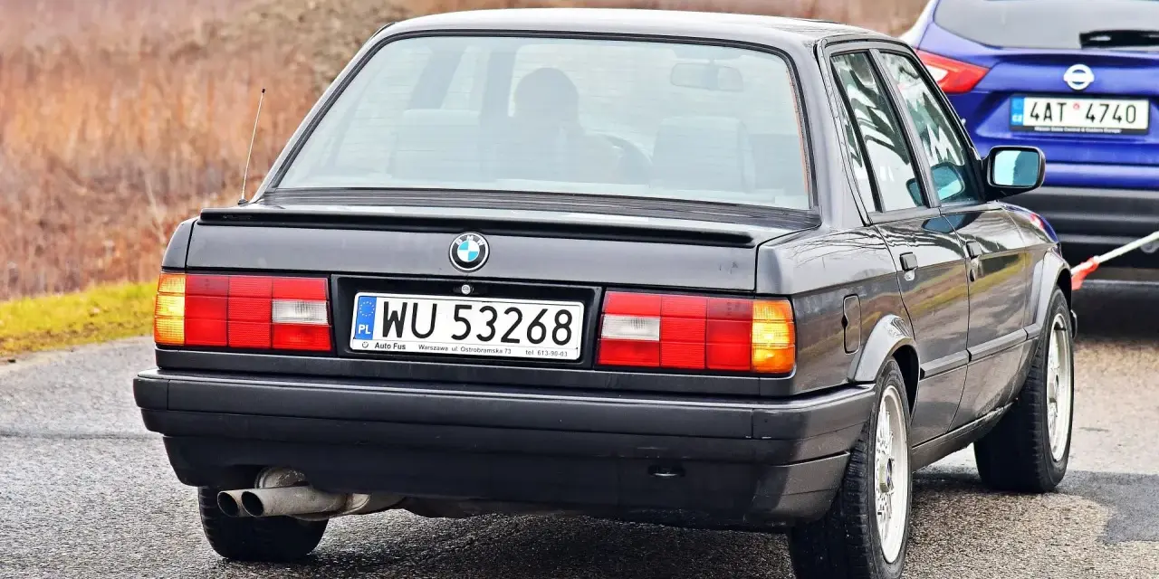 Tył czarnego BMW E30 z tablicą rejestracyjną WU 53268, holowanego przez niebieskiego SUV-a.