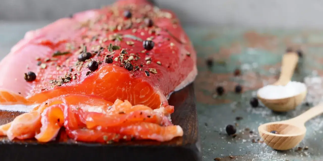 Un filet de gravlax de truite, assaisonné de poivre et d'herbes, prêt à être dégusté. Deux cuillères en bois contiennent sel et genièvre.
