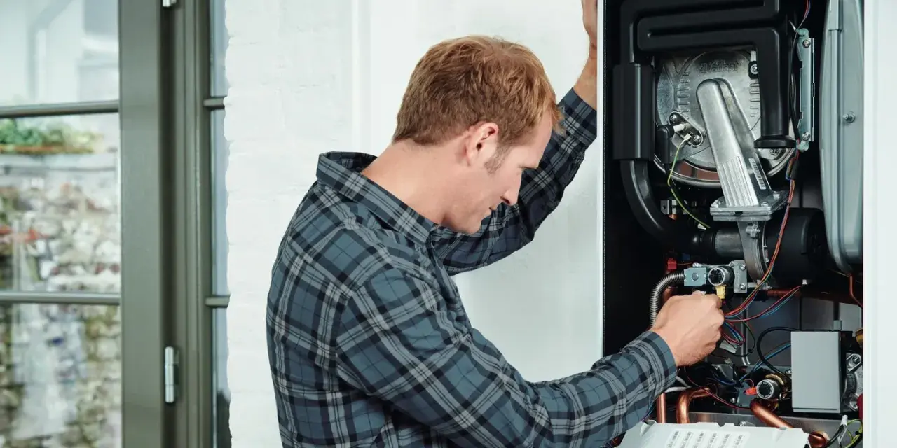 Tecnico che controlla lo scambiatore di calore della caldaia, con cavi e tubi visibili all'interno.