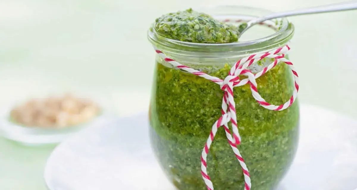 Un pot de pesto vert vif, décoré d'une ficelle rouge et blanche, prêt pour une recette pesto traditionnel. Quelques pignons de pin à côté.