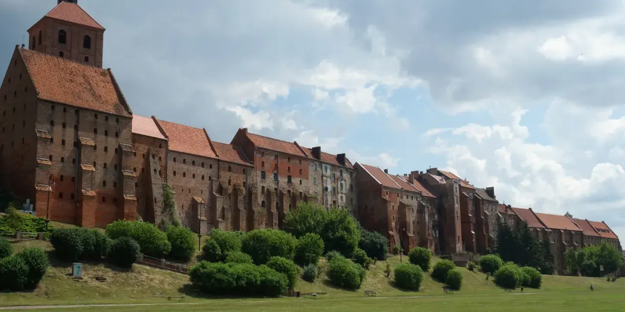 Długi, ceglany zamek w Grudziądzu w jeden dzień. Zielona trawa i krzewy przed zabytkową budowlą.