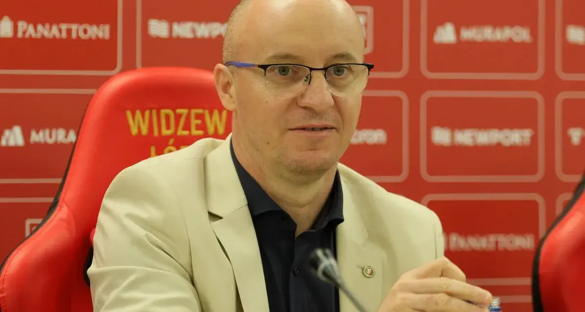 Nowy trener Widzewa w jasnej marynarce, siedzi na czerwonym fotelu z logo klubu.