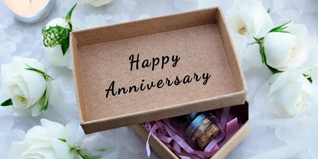 Otwarta szkatułka z napisem "Happy Anniversary" i małą buteleczką w środku, otoczona białymi różami i płonącą świecą. To idealny prezent na 4 rocznicę ślubu.