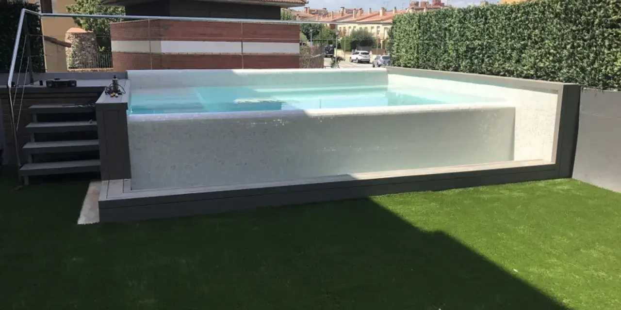 Moderna piscina con mosaico blanco, rodeada de césped artificial y setos. Ideal para decoración de terrazas con piscina.
