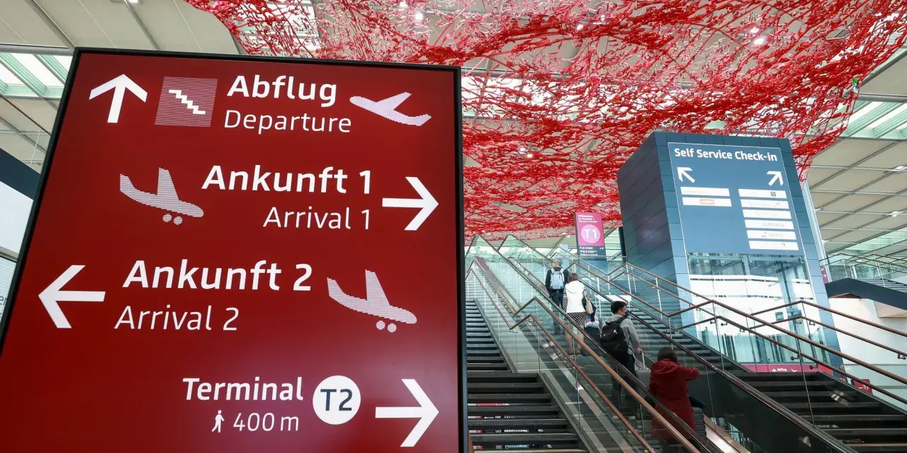 Czerwony znak z kierunkami na lotnisku w Niemczech: odloty, przyloty, terminal T2. W tle schody ruchome i instalacja artystyczna.