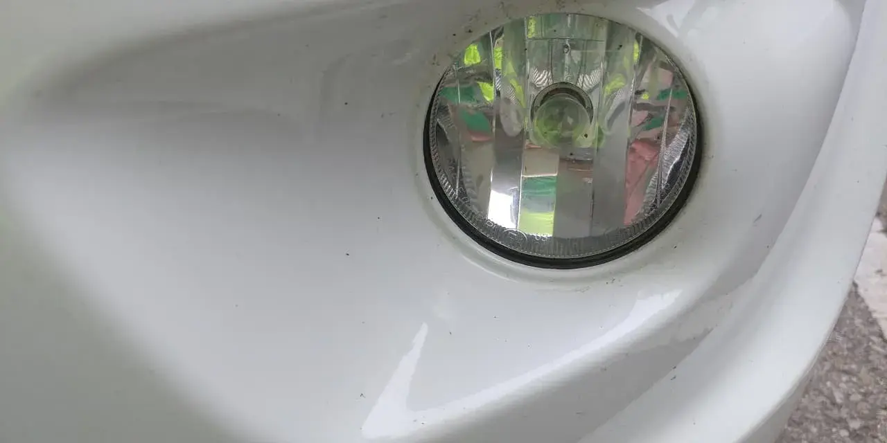 Zbliżenie na biały zderzak samochodu z widocznym okrągłym reflektorem. To światła przeciwmgielne przednie, lekko zakurzone, gotowe do jazdy w trudnych warunkach.